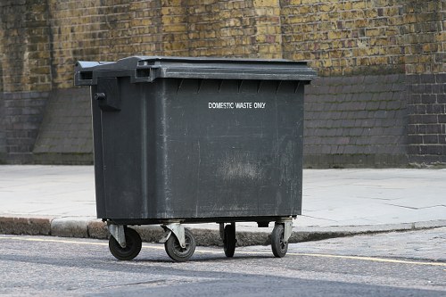 A large black plastic rubbish bin equipped with four small wheels is positioned on the pavement adjacent to a curb, with a rough textured brick wall and concrete pavement in the background. The bin's surface shows some signs of wear and dirt, and it features white text indicating it is for domestic waste only. The waste container has a hinged lid on top, which is closed, and is located on a street or alleyway, suggesting it is ready for waste collection services in Willesden. The surrounding environment includes a brick retaining wall behind the bin, and the pavement is comprised of concrete slabs with a slight slope. The scene is captured during daylight, providing natural, diffused lighting that highlights the texture of the brick wall and the surface of the bin. The image aligns with rubbish removal services offered by Commercial Waste Removal Willesden, illustrating the type of waste containers typically used in local residential or commercial areas within the London borough.
