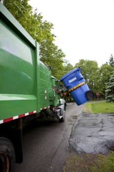 A large green waste collection truck parked on a paved residential street, with its rear open to reveal a compartment for rubbish. The truck is positioned adjacent to a blue wheelie bin that is partially lifted and tilted, indicating it is in the process of being emptied. The bin features a black lid, and a yellow strap is visible securing the lid or handle. Surrounding the scene, there are lush green trees and a grassy area on the right, with a driveway or pavement showing some wear and cracks. The environment suggests a typical suburban setting, possibly in or near Willesden, where Commercial Waste Removal Willesden provides rubbish removal services. The lighting indicates a cloudy day, with diffused natural light enhancing the clarity of the scene. The image captures the routine activity of waste collection, emphasizing the importance of professional rubbish removal in maintaining local cleanliness and environmental standards.