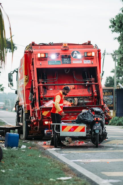 A worker in a high-visibility yellow and orange vest is standing beside an open rear compartment of a large red rubbish collection truck, which is parked on the side of a street. The truck has a metal