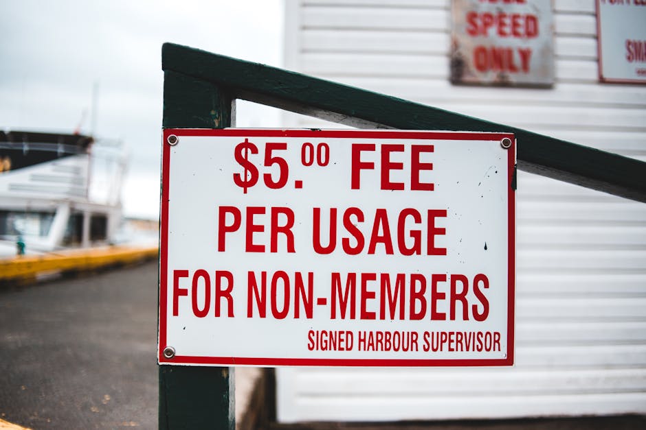 A rectangular white sign with bold red lettering displaying a fee of five pounds per usage for non-members, signed by the harbour supervisor, is mounted on a dark green metal post placed beside a pave